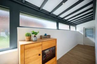 veranda_surface.webp Un couloir lumineux avec un plafond en poutres apparentes et un meuble en bois décoré de plantes, baigné de lumière naturelle par des fenêtres