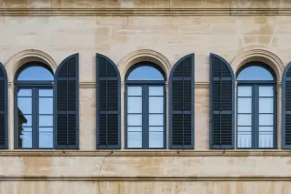 volet_aluminium.webp Façade d'un bâtiment classique avec des fenêtres arquées et des volets noirs contrastant sur un mur de pierre beige.