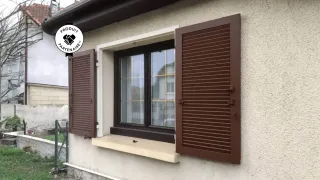 Fenêtre avec volets marron ouverts sur une maison beige, arborant un badge 'Produit Partenaire'.