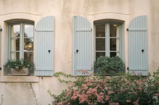volet_bois.webp Façade de maison provençale avec des volets bleus et des plantes fleuries en avant-plan.