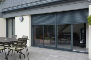 volet_isolation_acoustique.webp Une terrasse moderne avec des chaises et une table en métal, adossée à une maison équipée de volets roulants gris sur de grandes ba