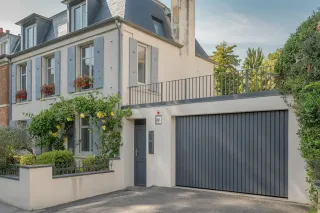 volet_isolation_phonique_intret.webp Maison de style moderne avec façade crépie, volets gris, garage intégré et jardin fleuri.