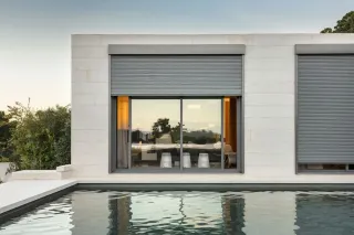 Volet_maison.webp Vue extérieure d'une maison moderne avec des volets roulants fermés et une piscine à l'avant, reflétant le paysage environnant.