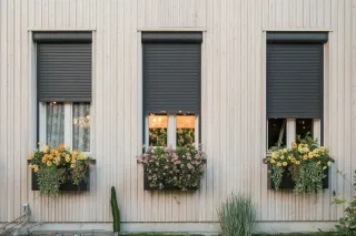 volet_pvc.webp Trois fenêtres ornées de volets roulants gris foncé et de jardinières fleuries décorent la façade en bois clair d'une maison