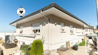 Une maison moderne et lumineuse avec des volets roulants fermés, entourée d'un jardin entretenu sous un ciel bleu clair.