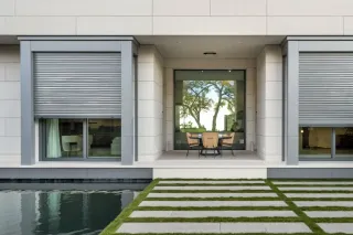 volet_roulant_intgrs.webp Façade moderne d'une maison avec un patio élégant, des stores métalliques, et une vue sur un jardin avec une piscine.