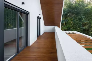 volet_roulant_isolation_acoustique.webp Une terrasse moderne en bois avec une balustrade blanche et de grandes baies vitrées, offrant une vue sur la nature environnante.