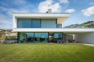 volet_roulant_isolation_phonique.webp Vue d'une maison moderne à deux étages avec de grandes baies vitrées, entourée d'un jardin verdoyant sous un ciel bleu.