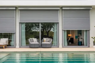 volet_roulant.webp Un salon moderne avec de larges baies vitrées dotées de volets roulants, donnant sur une piscine et un jardin arboré.