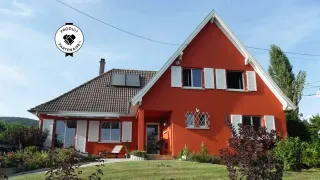 Maison moderne avec une façade orange et des volets blancs, entourée d'un jardin verdoyant sous un ciel bleu clair.