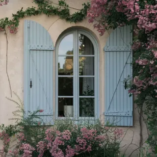 volets_battants.webp Fenêtre avec volets bleus encadrée de fleurs roses et de verdure sur une façade en pierre.