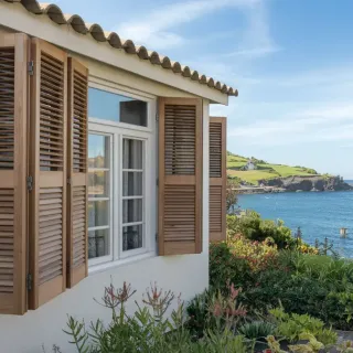 volets_pliants.webp Vue d'une maison en bord de mer avec des volets en bois, entourée de végétation verdoyante et offrant un magnifique panorama sur l