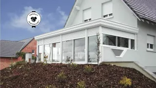 Une maison moderne avec une grande véranda vitrée, située sur une colline paysagée, sous un ciel ensoleillé.